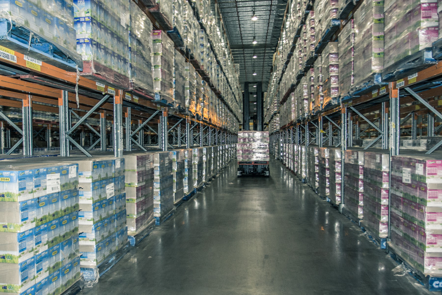 Wide warehouse aisle with tall racks and forklift moving boxed goods in the center.