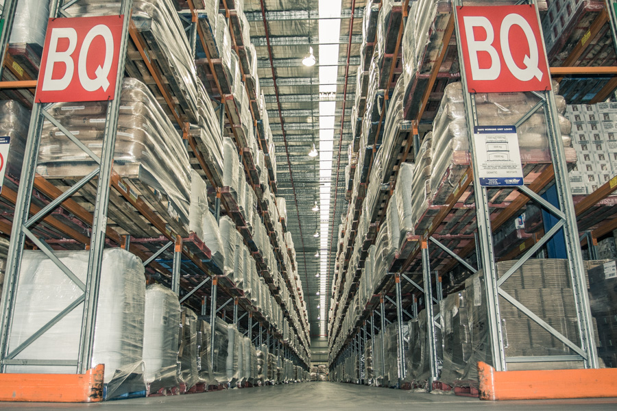 Wide warehouse aisle with high racks, wrapped pallets, and large red "BQ" signs.