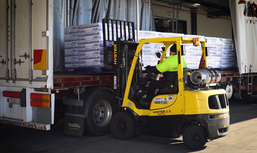 Forklift operator in neon yellow jacket loads pallets onto truck amid shipping equipment.