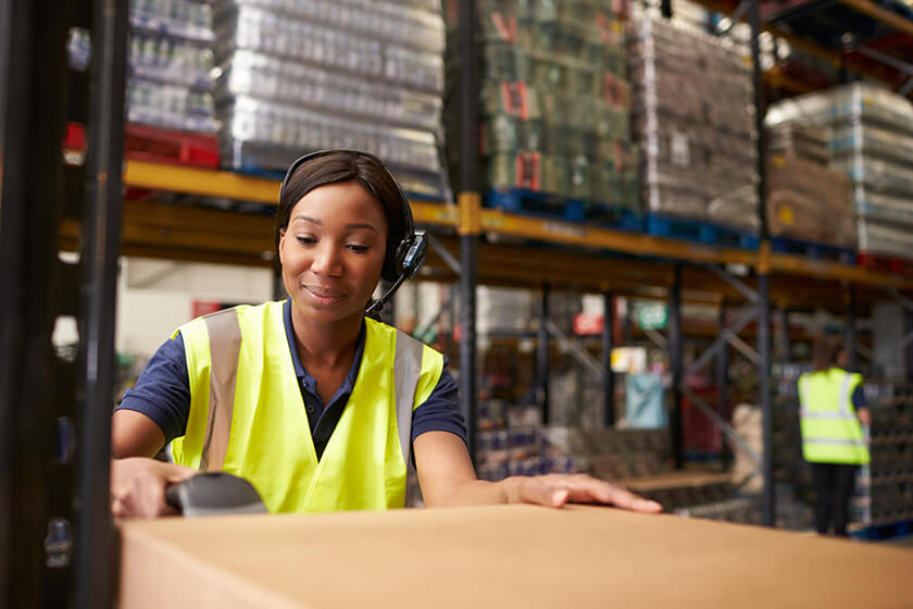 Warehouse worker in bright vest operates machinery, focused amid shelves and stacked goods.