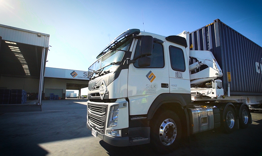 White logistics truck with crane parked by "SILK Contract Logistics" warehouse under blue sky.