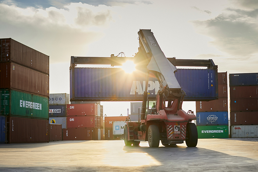 Reach stacker lifts container at sunset in busy yard with colorful stacked containers.