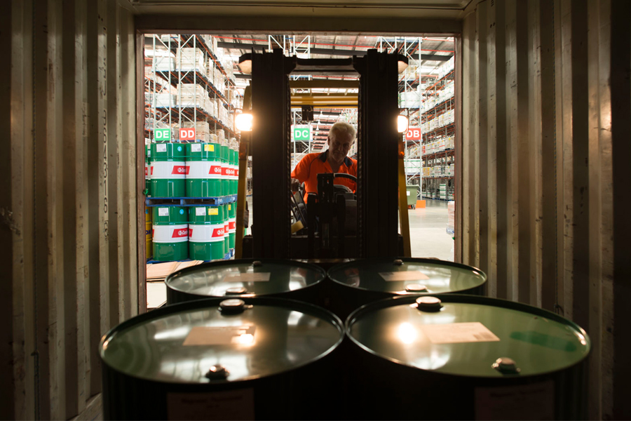 Forklift operator inside shipping container with green barrels and industrial shelving behind.